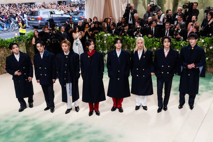 Group of individuals in matching dark coats and various hairstyles standing in a row, likely a music band