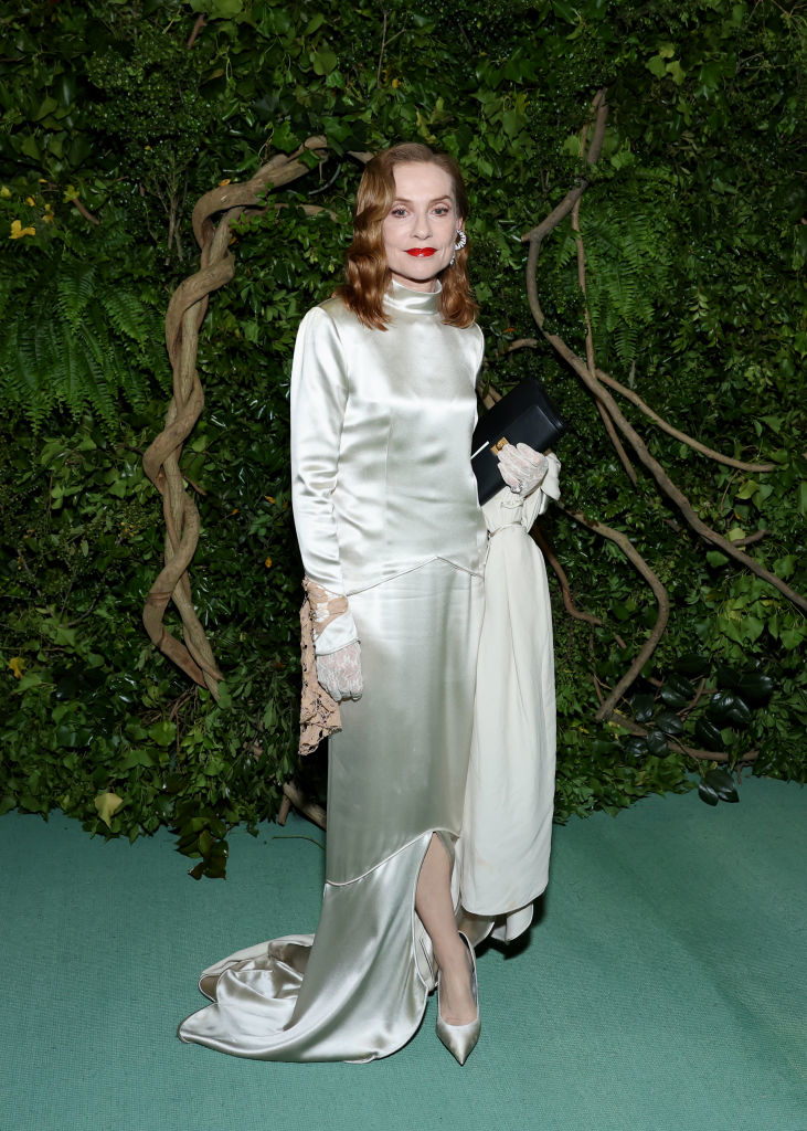Isabelle Huppert on greenery backdrop wears elegant satin gown with gloves, holding a clutch