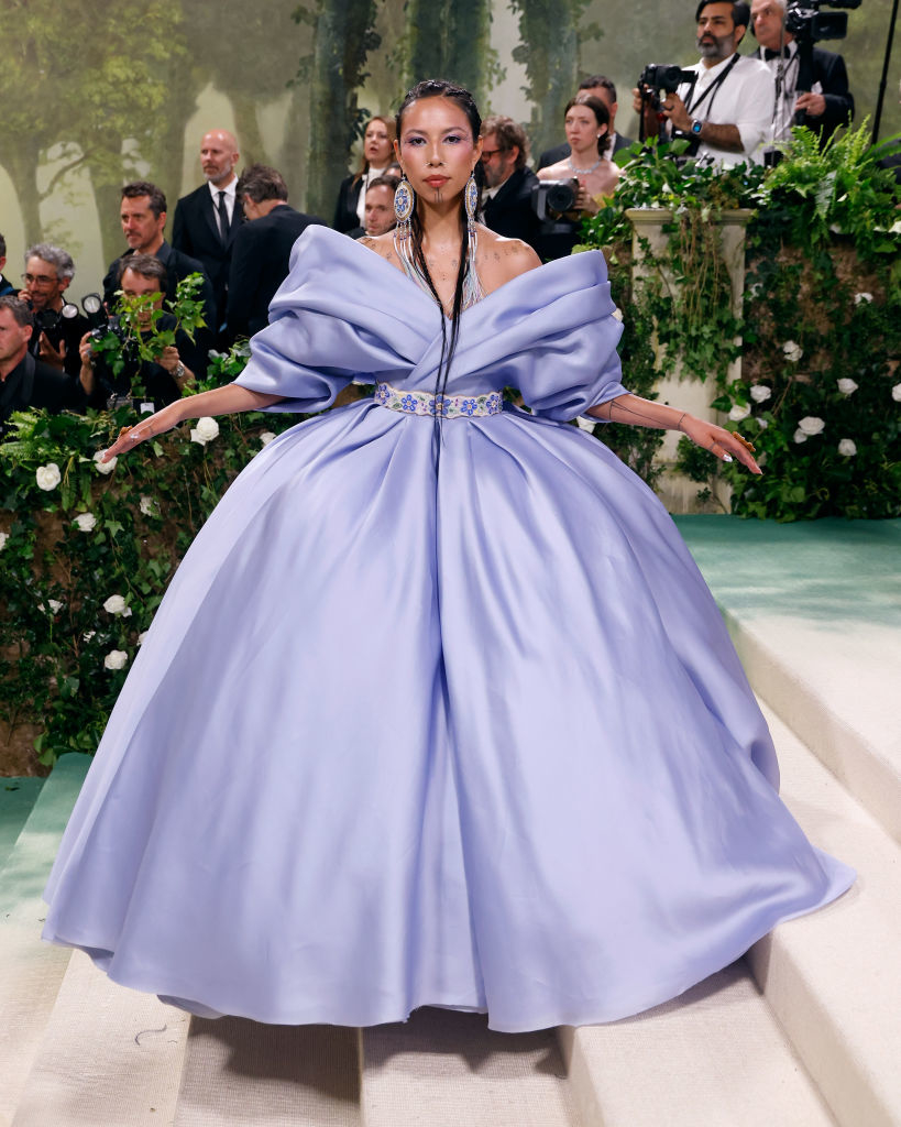 Quannah Chasinghorse in a voluminous off-the-shoulder lilac gown with a cinched waist detail, against a floral backdrop