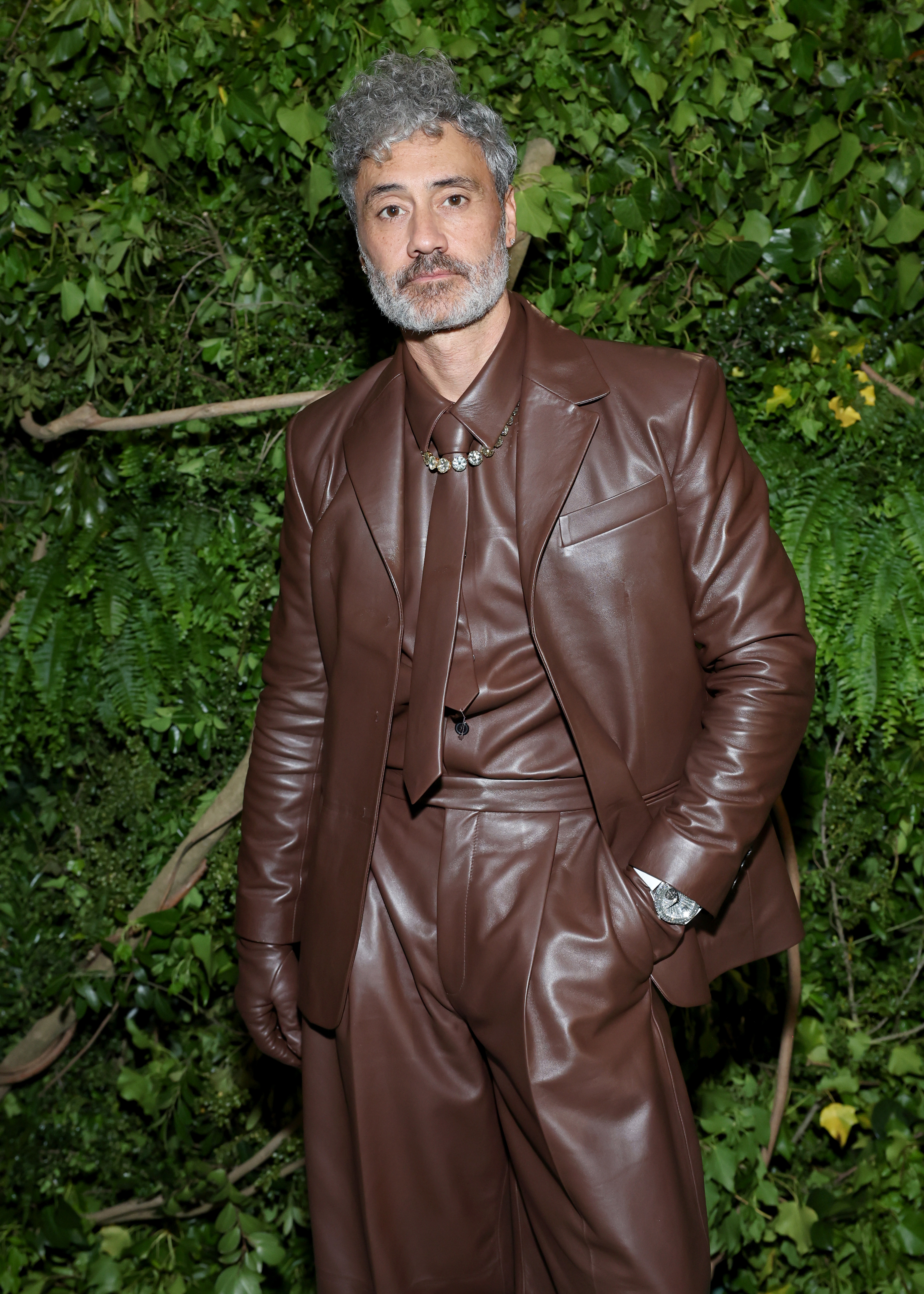 Taika Waititi in a stylish leather outfit with a statement necklace poses against a leafy backdrop