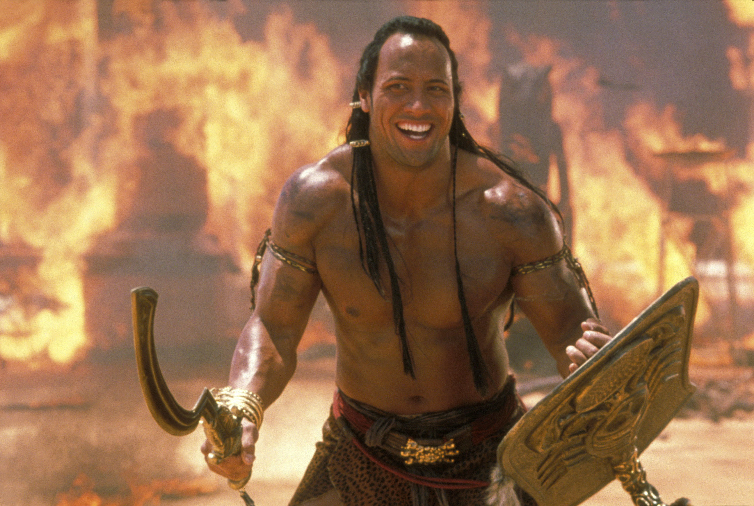 Dwayne Johnson in a warrior costume with a sword and shield smiling in front of a blazing backdrop