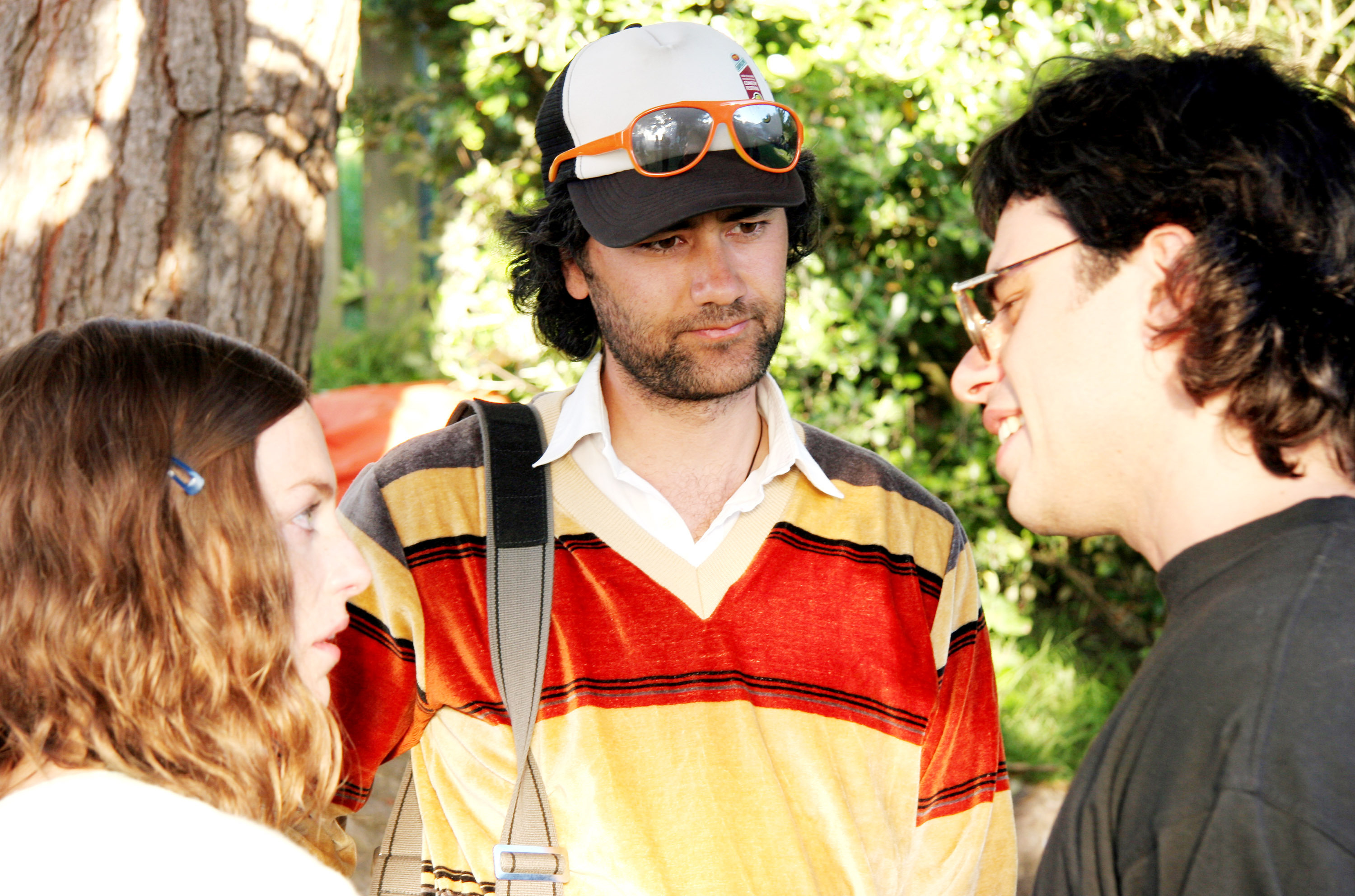 Loren Horsley, director Taika Cohen and Jemaine Clement engaging in conversation outdoors, one wearing a striped shirt and cap with sunglasses