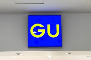 Sign with "GU" logo on a blue background, mounted on a wall with ceiling lights visible
