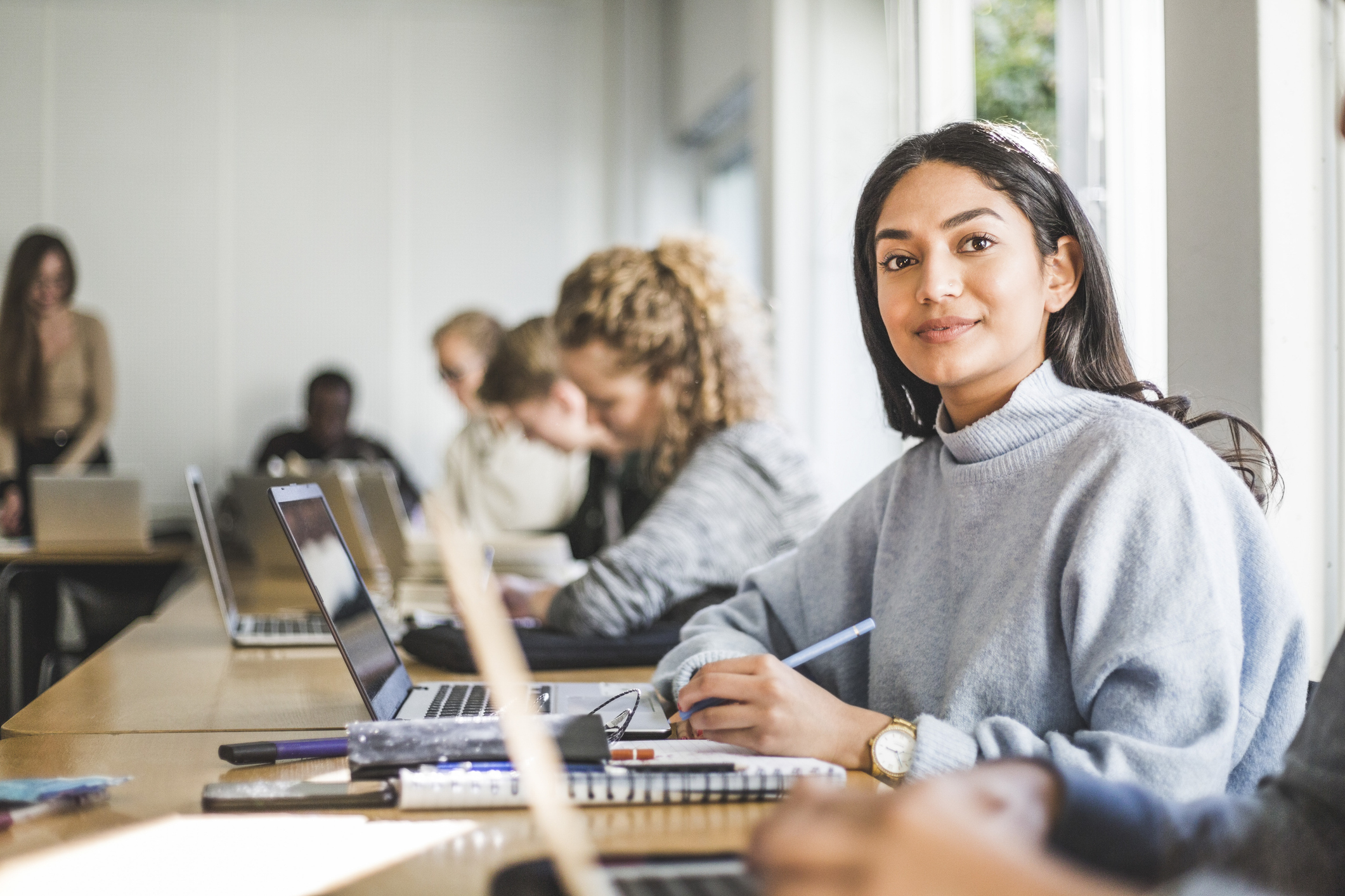 Woman smiling at desk among other people working on laptops in a classroom or office setting