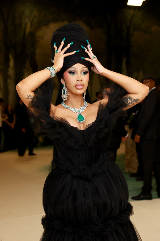Cardi B in extravagant black gown with large gemstone jewelry, posing with hands on headwear at an event