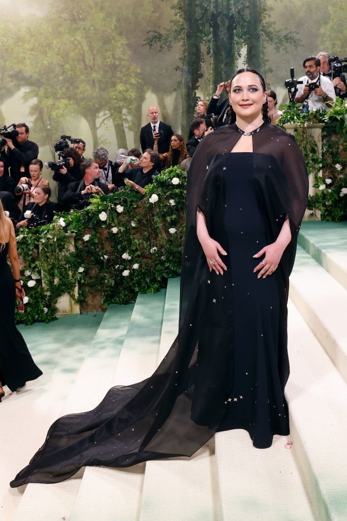 Lily Gladstone in an elegant black gown with cape at an event, photographers in background