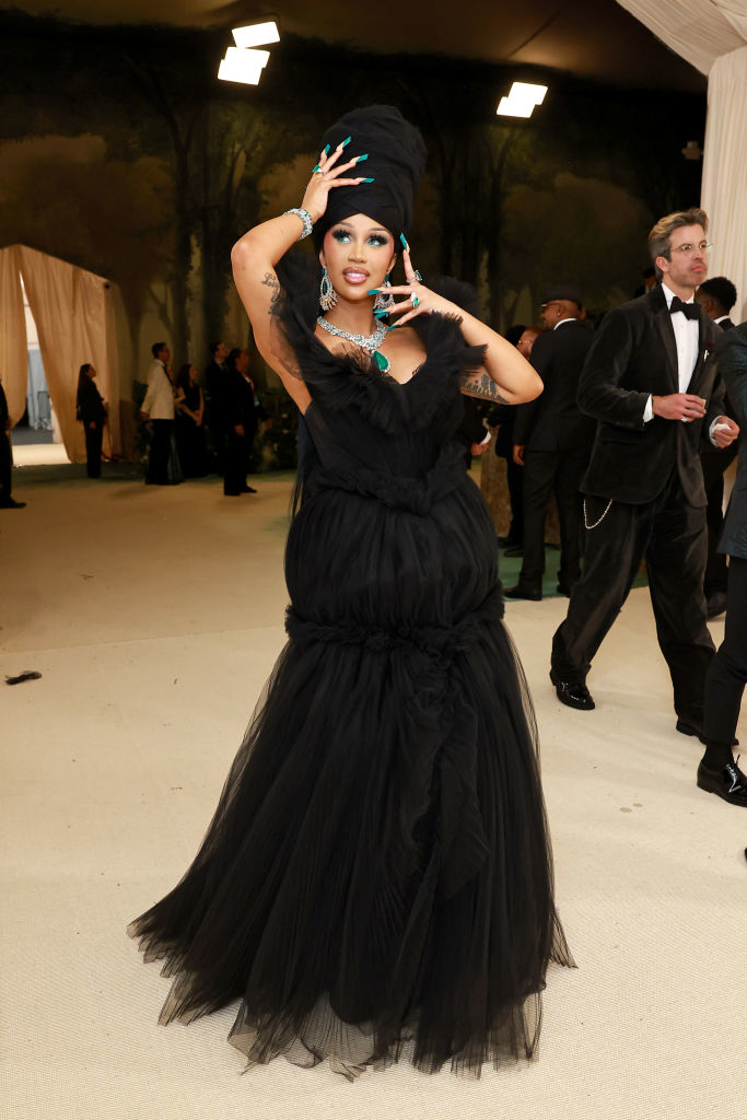 Cardi B in a voluminous black gown with elaborate headpiece, posing with hand on head at an event
