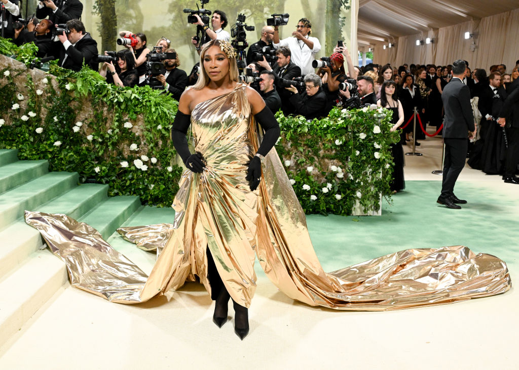 Serena Williams in a shiny, draped gown with a long train at an event, posing for photographers