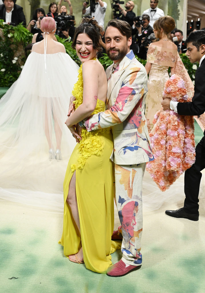Two celebrities at an event, woman in yellow gown with floral details, man in patterned suit with artistic print