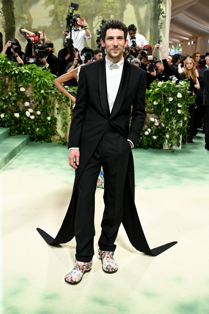 Man in a black tuxedo with unique long-tail design and patterned shoes stands on event carpeting; photographers in background