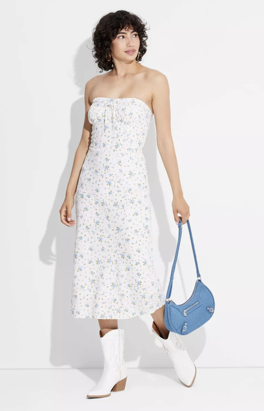 person in a strapless floral dress with a blue bag and white boots posing against a plain background