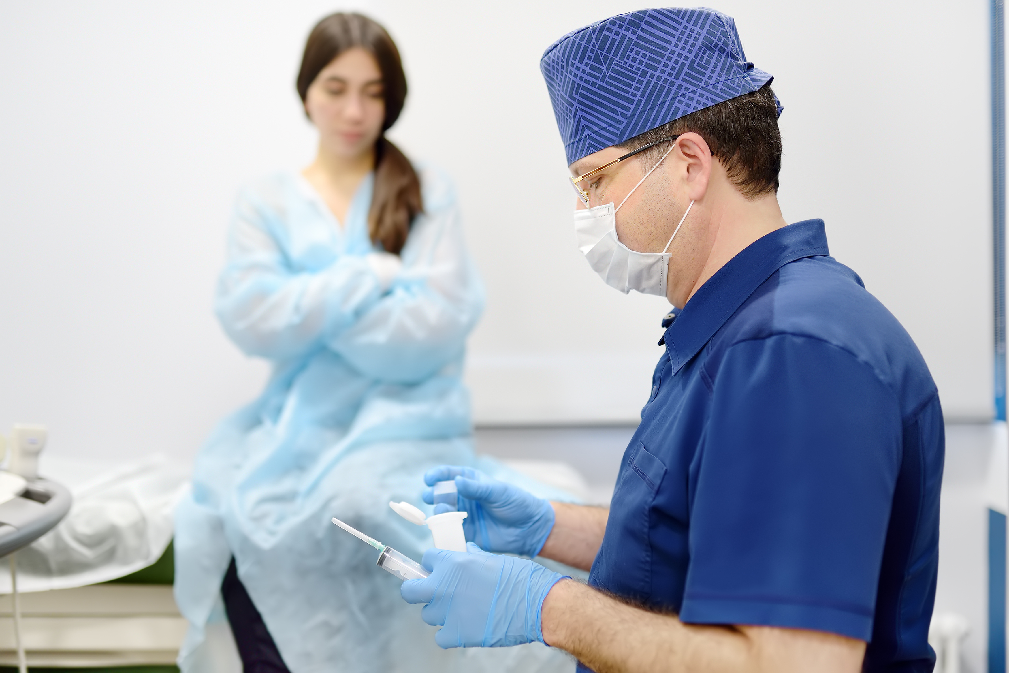A doctor in scrubs prepares a syringe as a patient sits on the table