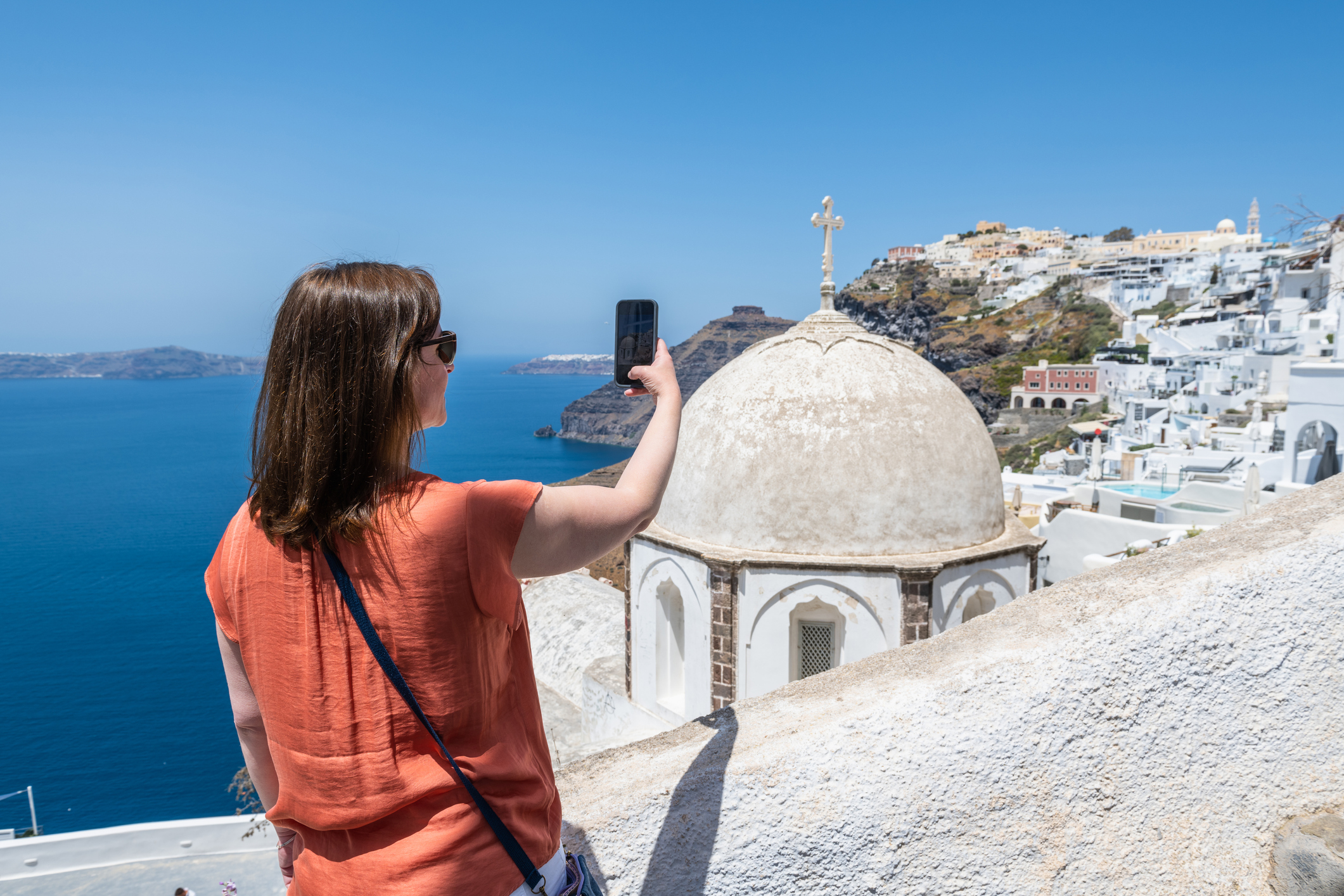 A person taking a selfie with a smartphone against the backdrop of a coastal town with distinctive architecture