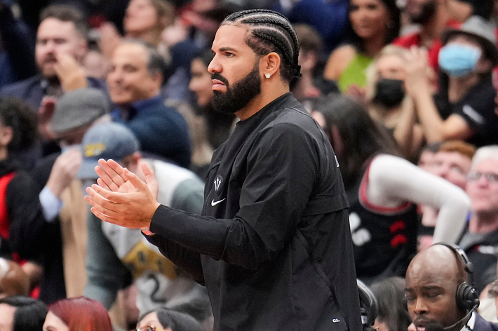 Drake in a black outfit clapping at a basketball game from the sidelines