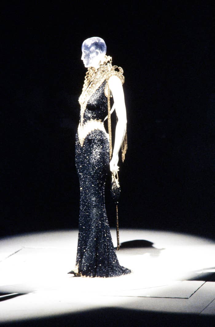 Model on runway in a fitted, glittery gown with an ornate shoulder piece and a moon headpiece