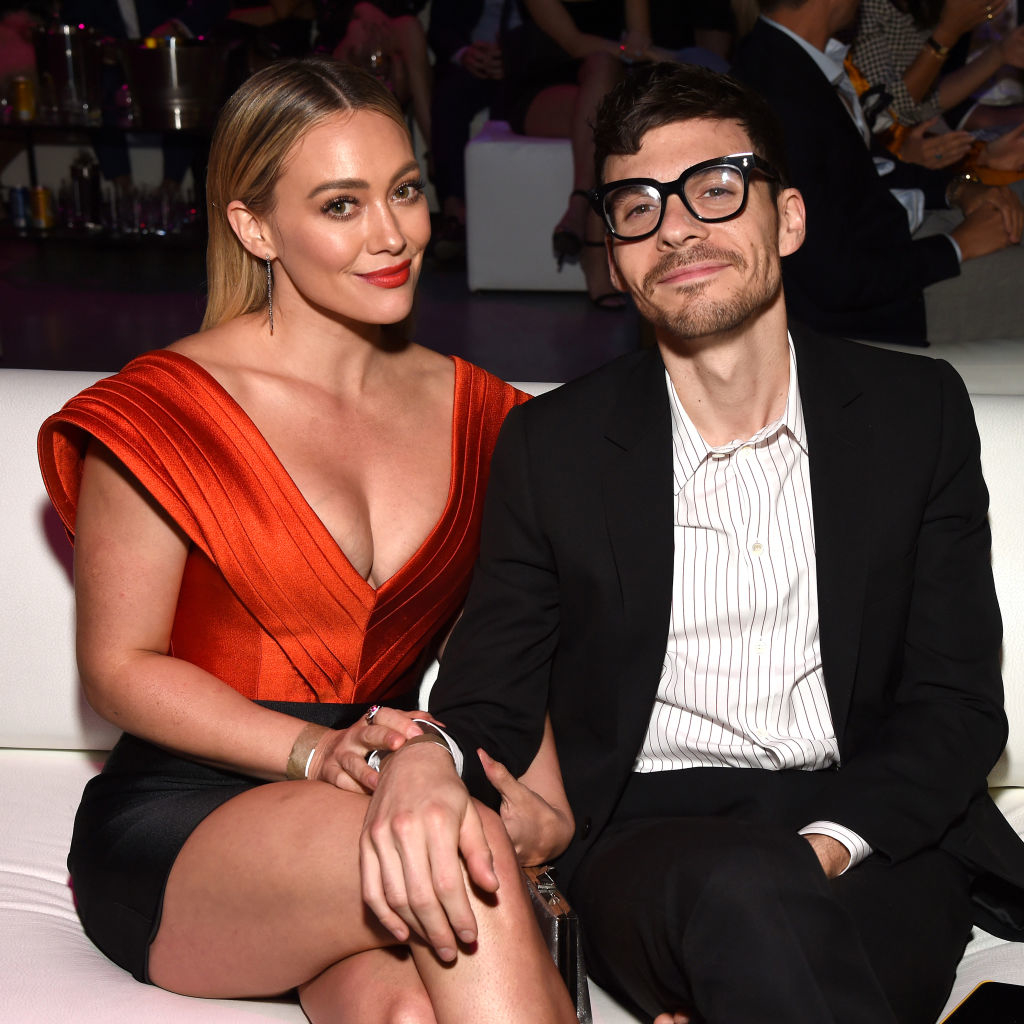 Hilary Duff in a red V-neck dress seated next to Matthew Koma in a striped suit at an event