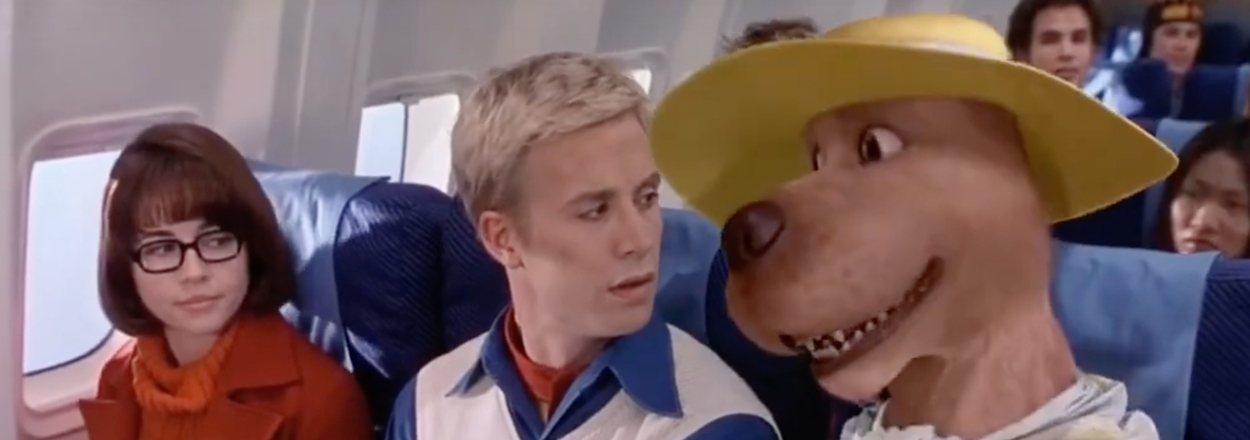 Velma, Fred, and Scooby-Doo, in airplane seats, Scooby wearing a large hat and scarf