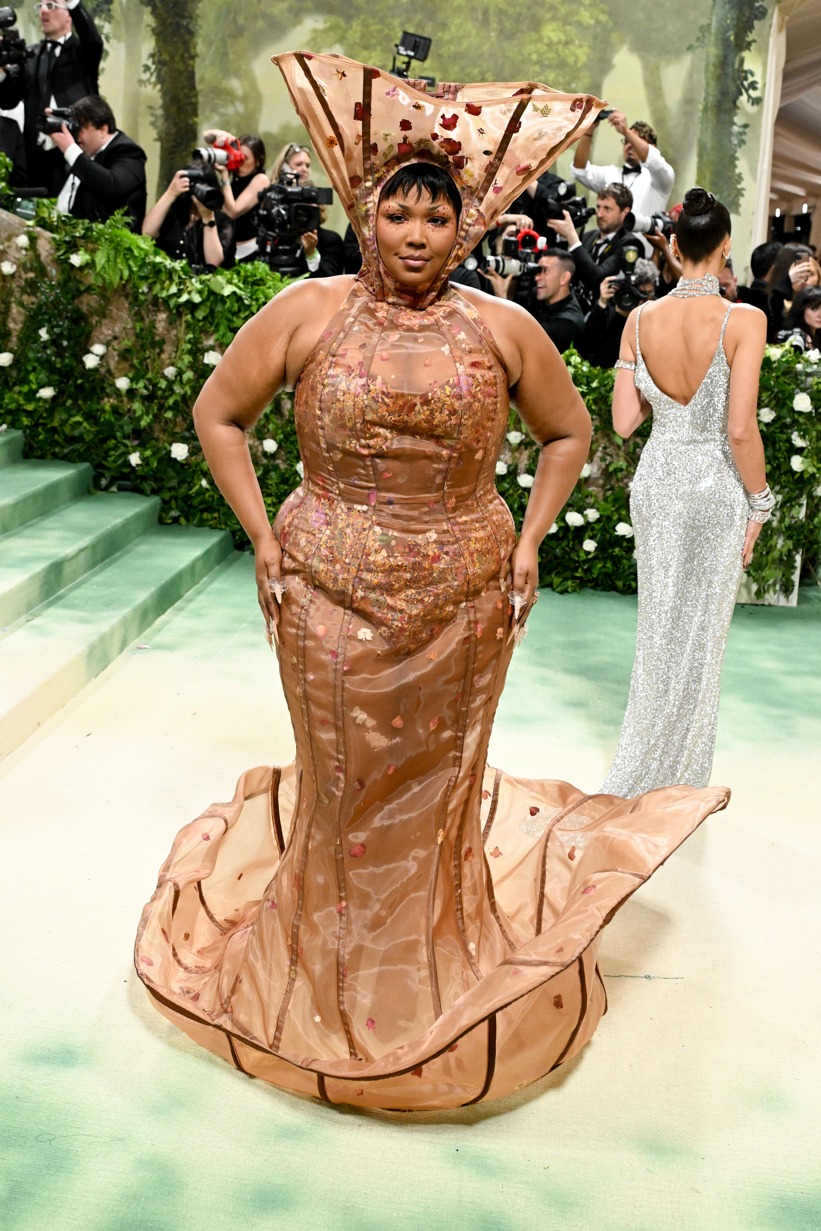 Lizzo in a flamboyant gown with oversized ruffled collar, posing on stairs at an event. Behind her, another guest in a sleek gown