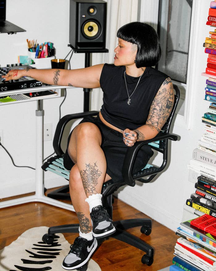 Woman in a black dress and sneakers, seated by a desk, surrounded by books and speakers