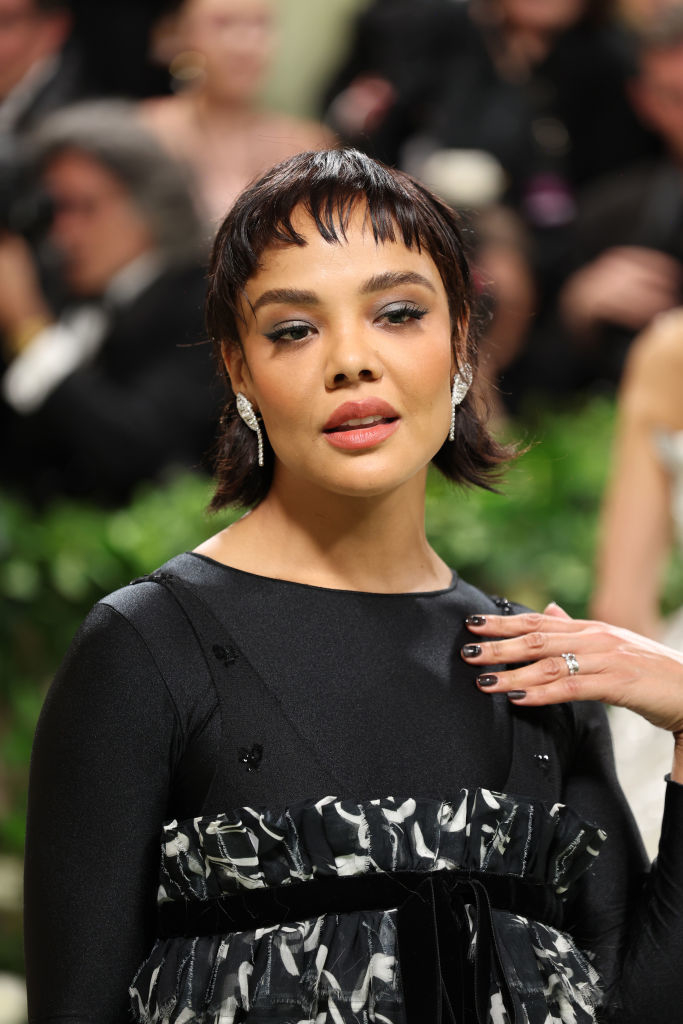 Tessa Thompson with bob haircut and embellished black dress poses at an event, wearing earrings and a ring