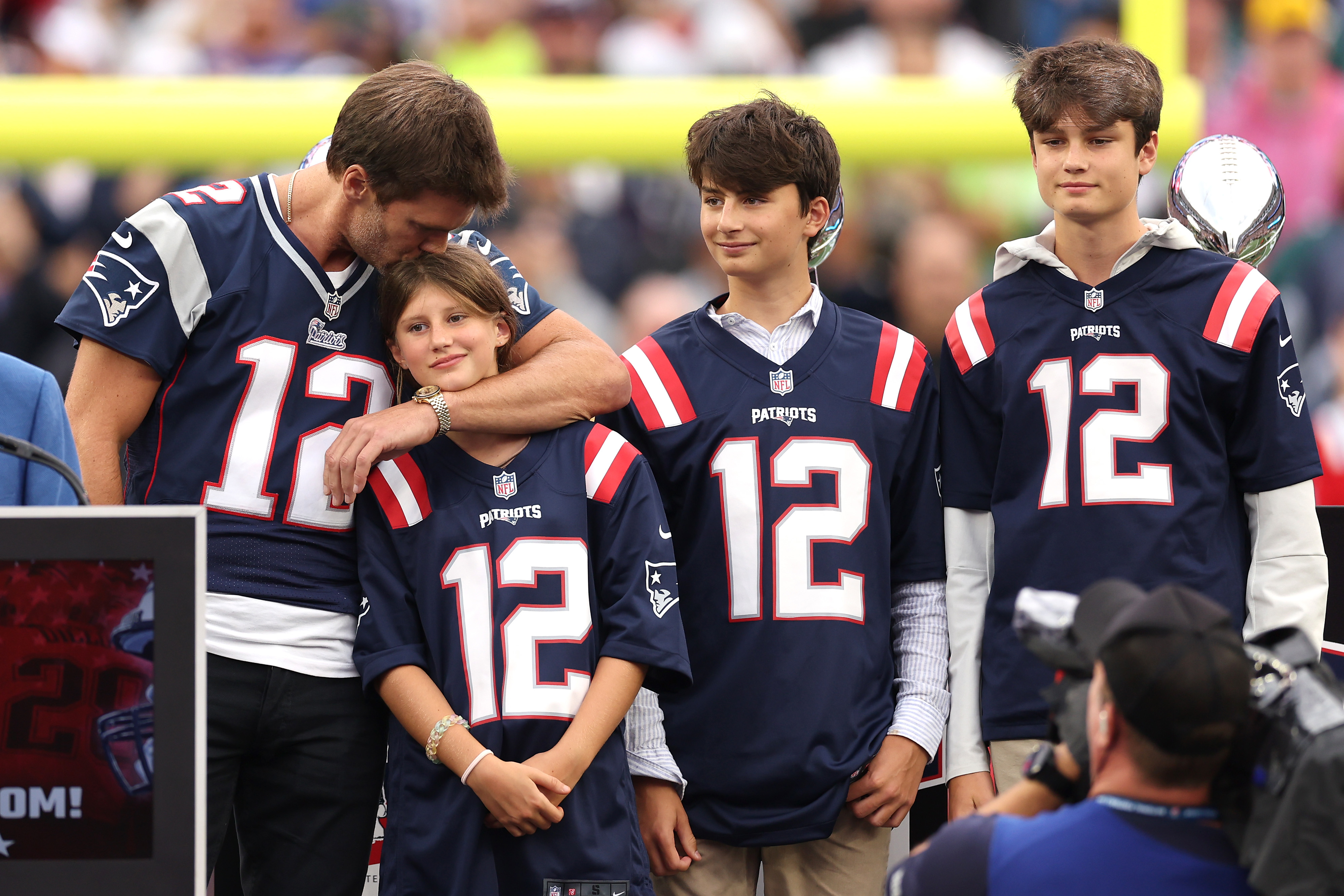 Tom Brady hugging a child, with two others beside him, all in Patriots jerseys at a stadium event
