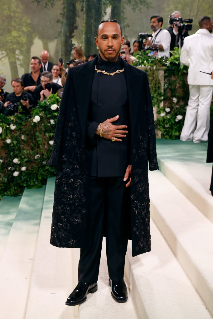Lewis in a luxurious black embroidered coat and slacks on a carpet, photographers in the background