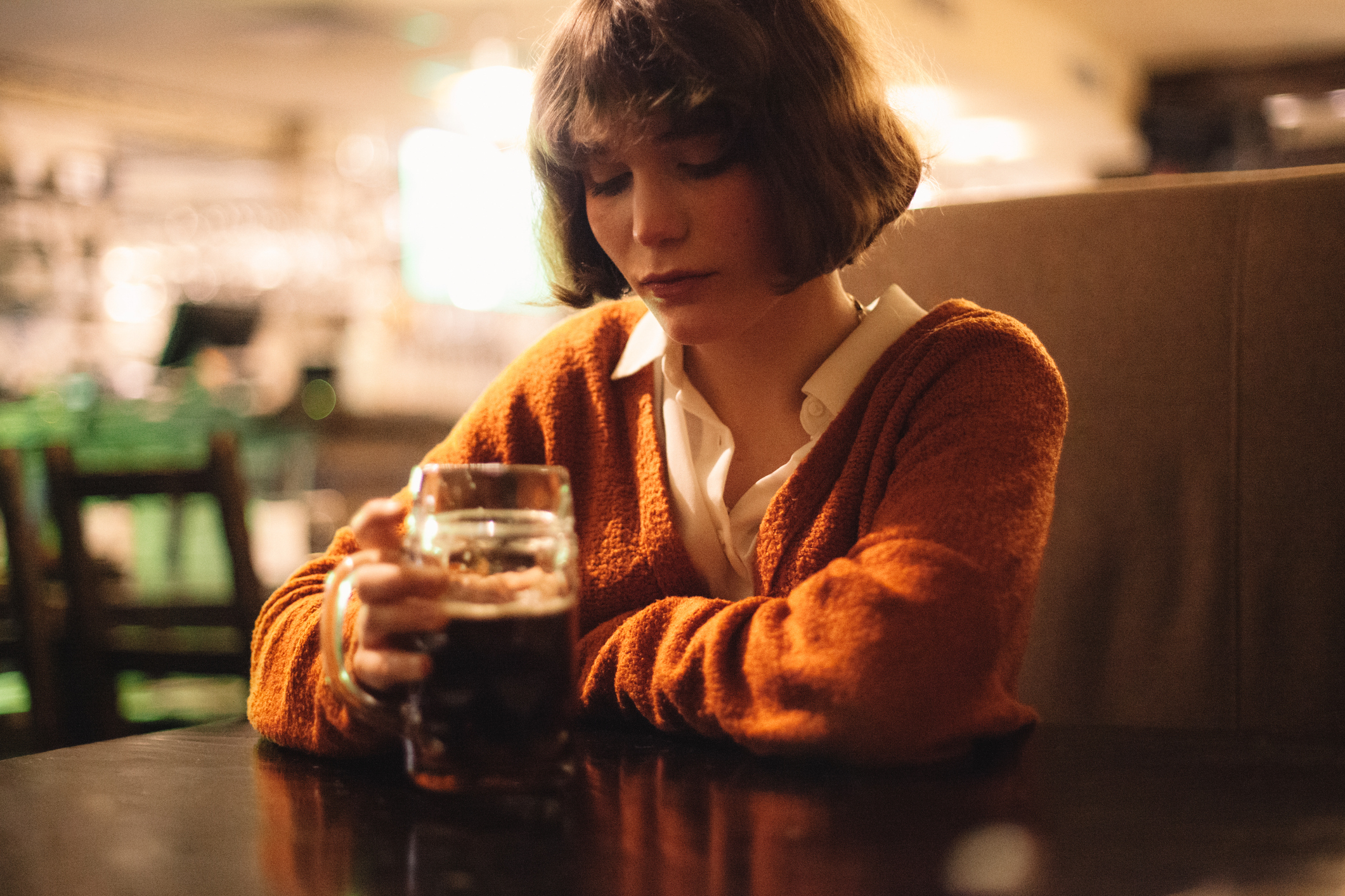 Woman in an orange cardigan holding a drink at a table, with contemplative expression