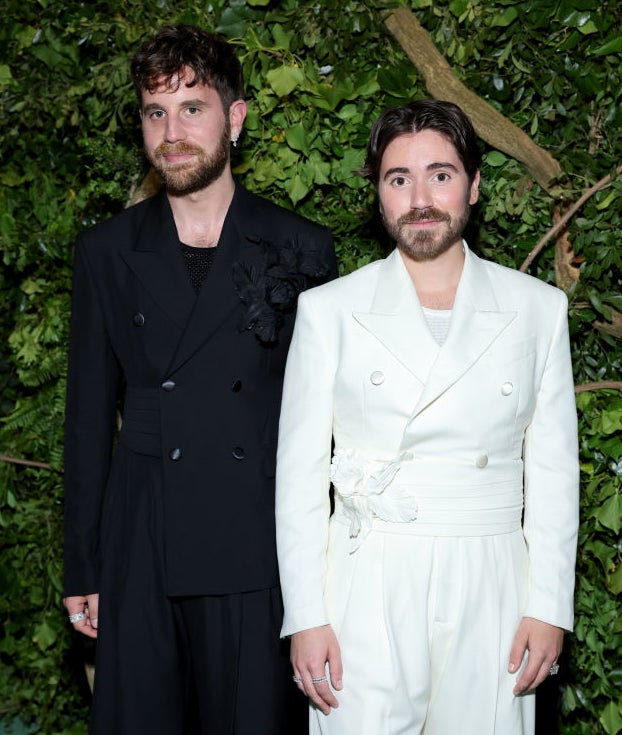 Ben Platt and Noah Galvin on greenery backdrop; one in a black suit with floral detail, the other in white tied jacket and pants