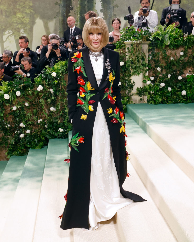 Anna Wintour in a long black coat with bright floral embroidery, standing at an event with photographers in the background