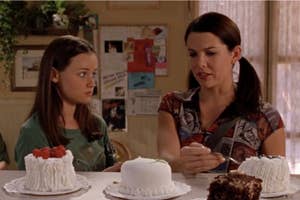Rory and Lorelai from Gilmore Girls sit at a table looking at each other with cakes in front of them