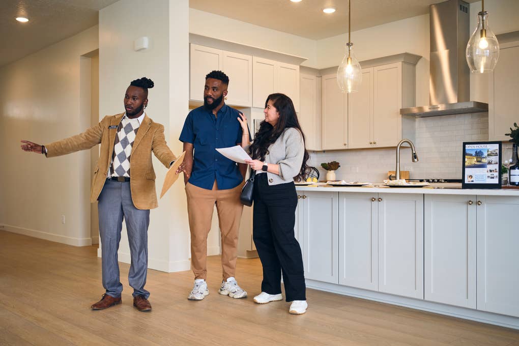 Realtor showing a kitchen to a couple, gesturing while they hold documents and listen