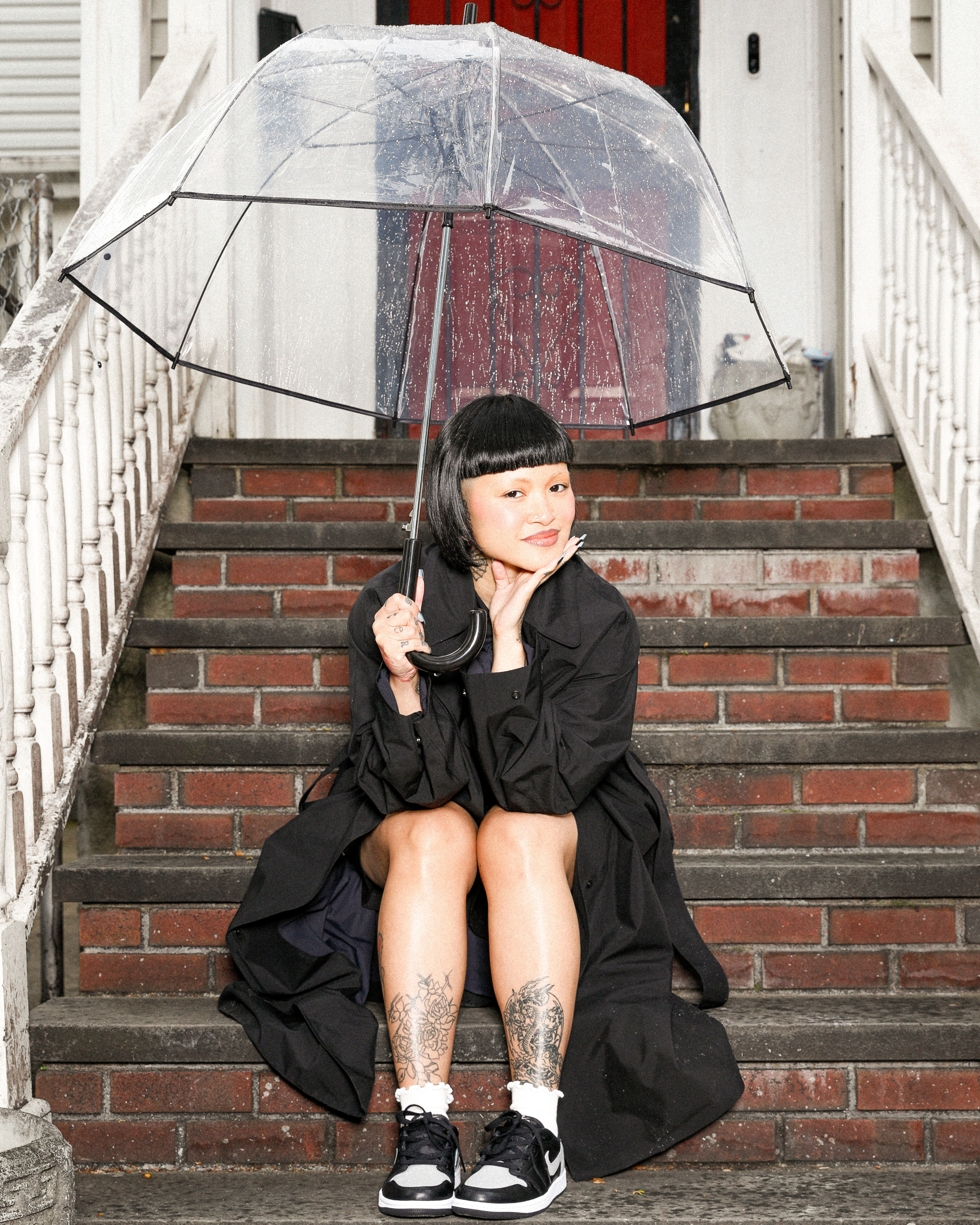 Person sitting on steps under a clear umbrella, wearing a black coat and patterned sneakers