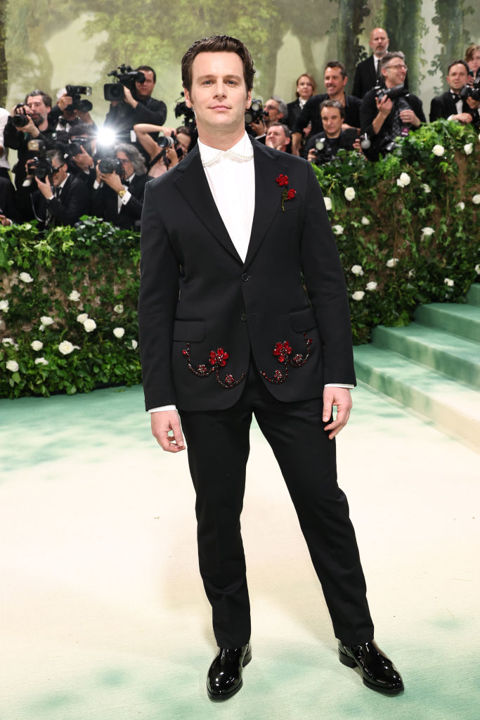Jonathan Groff in a black suit with floral embroidery on the jacket, standing on a stairway