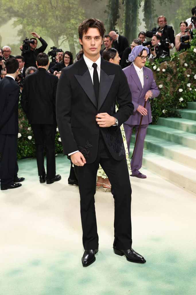 Nicholas Galitzine in black suit with embroidered details stands on stairs; photographers in background
