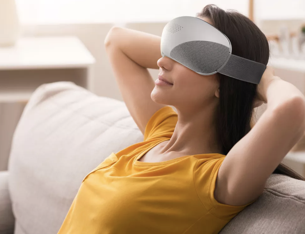 Model relaxing with a gray sleep mask on a sofa, hands behind head, in casual attire.