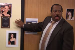 Stanely from "The Office" in a business suit gesturing to a wall with framed photos of Hilary Swank