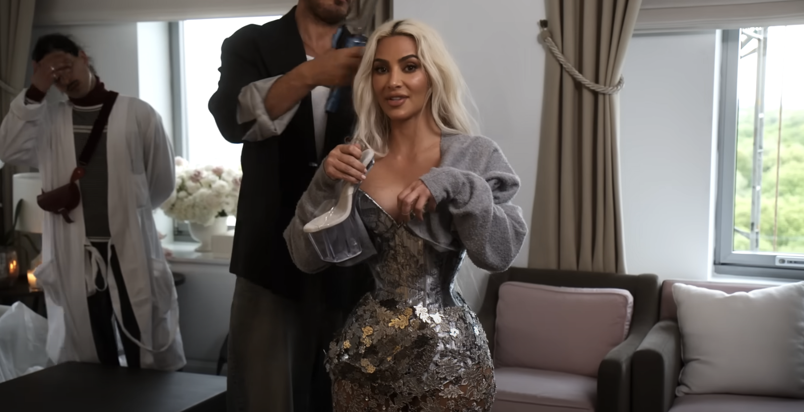 Kim Kardashian in a glittery fitted dress holding a glass while a stylist adjusts her hair
