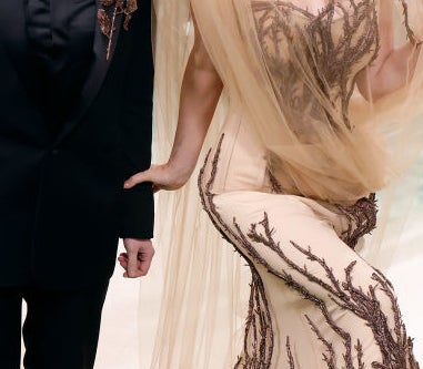 Two people on event carpet, one in black suit with floral embroidery, the other in sheer gown with branch-like design