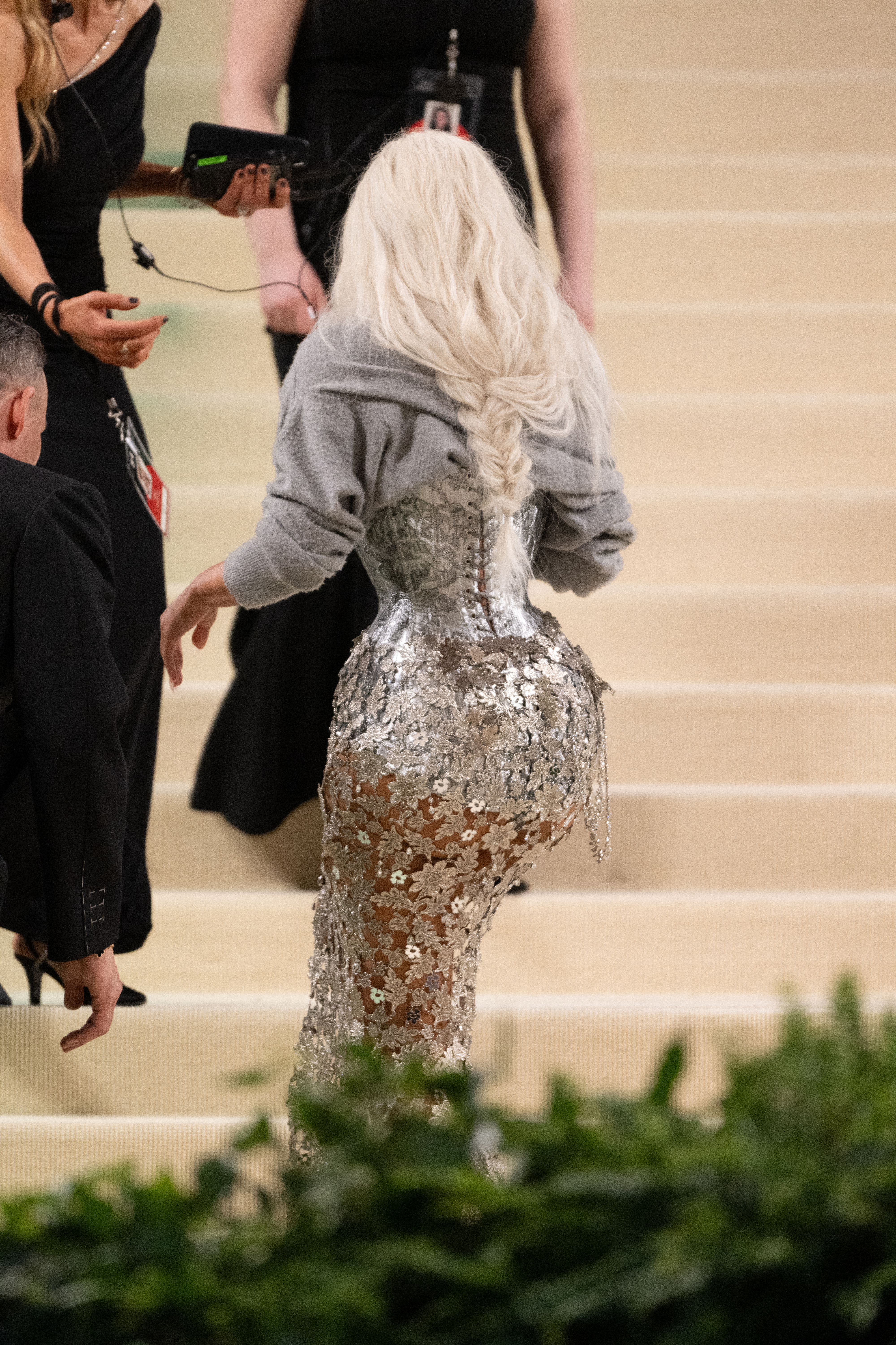Person on red carpet in sparkling silver dress with large bow detail, viewed from behind. Hair styled in long waves