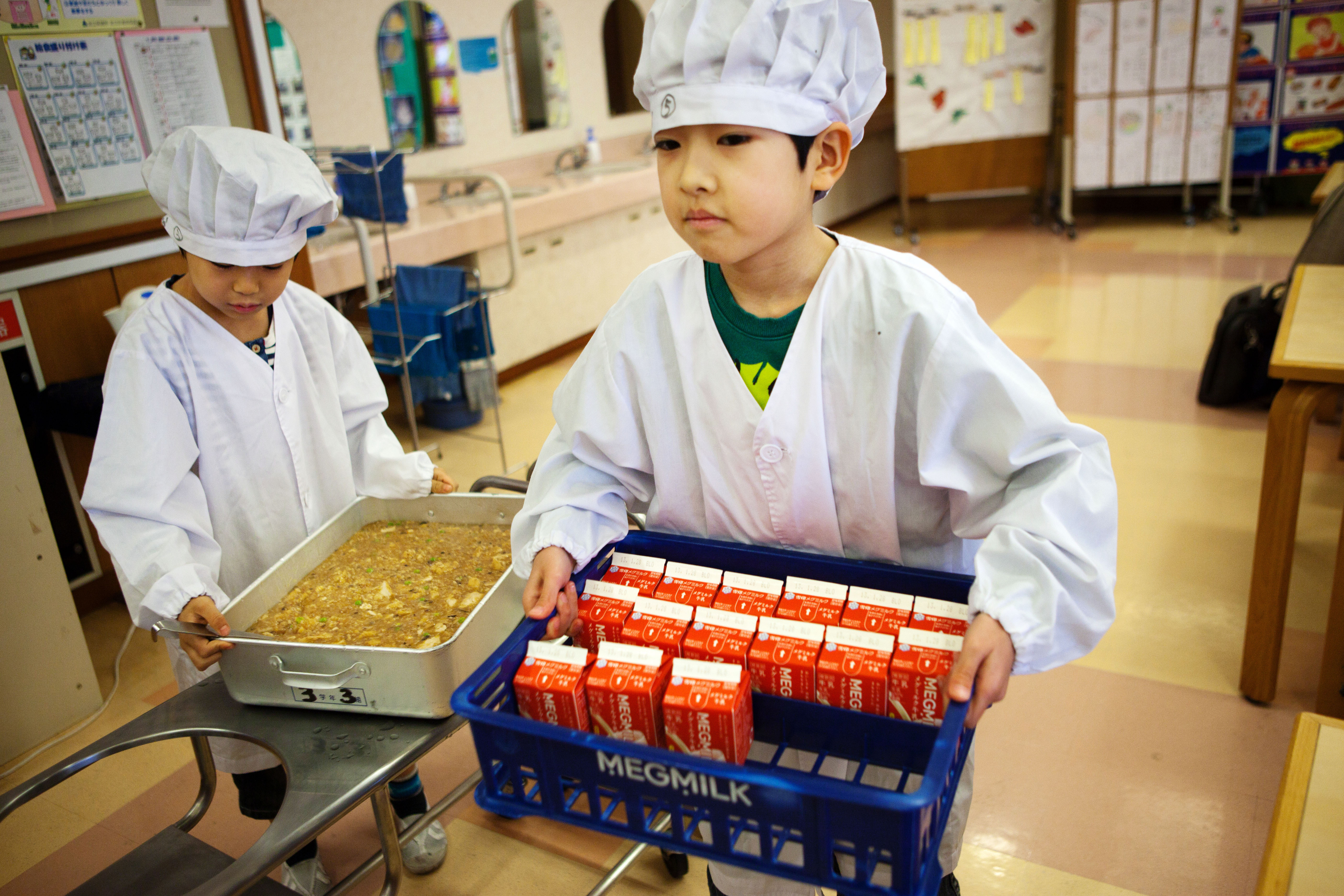 Two children in chef hats and aprons are serving milk cartons at a school