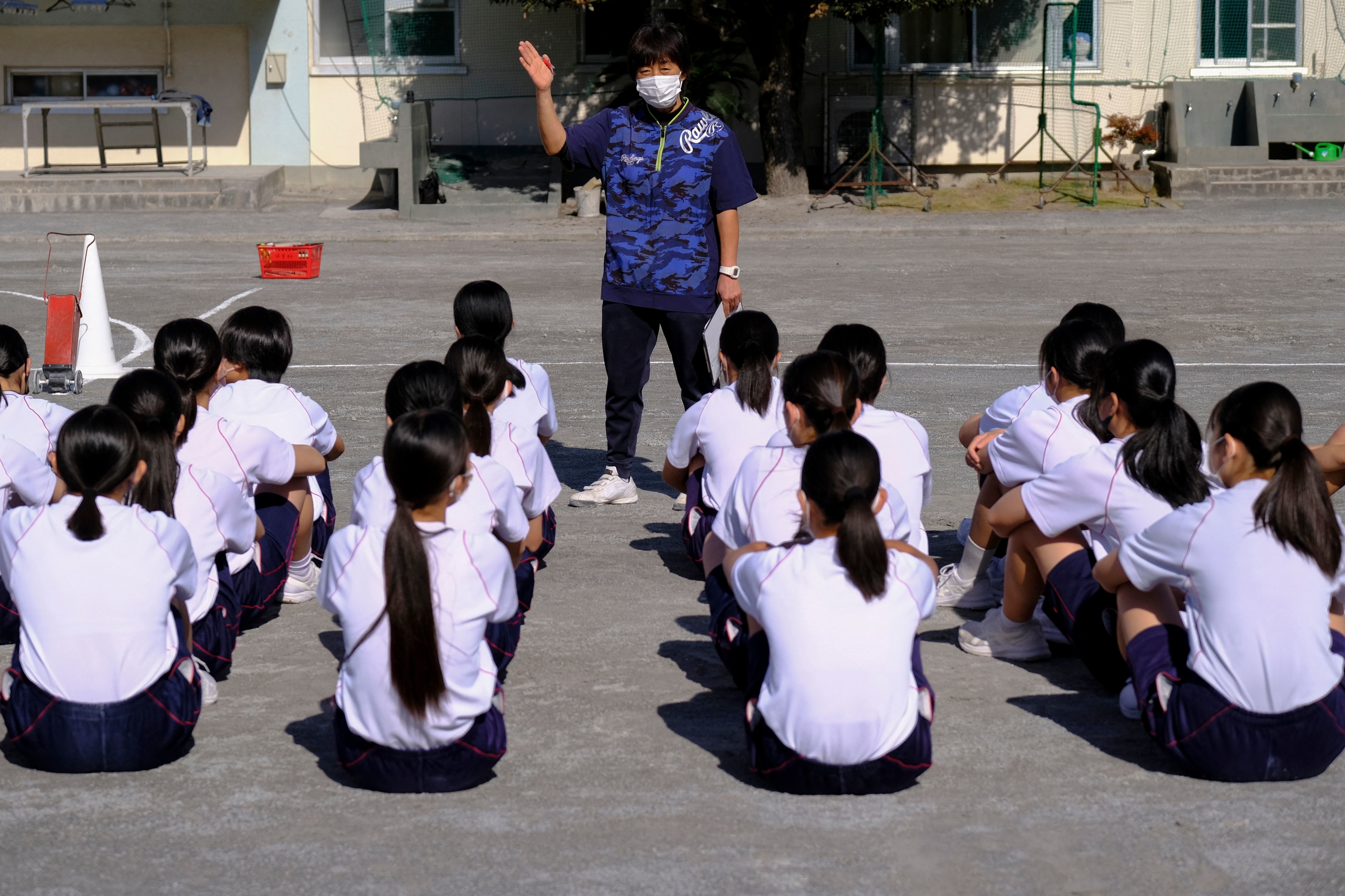 Instructor with face mask standing before seated students in schoolyard, gesturing