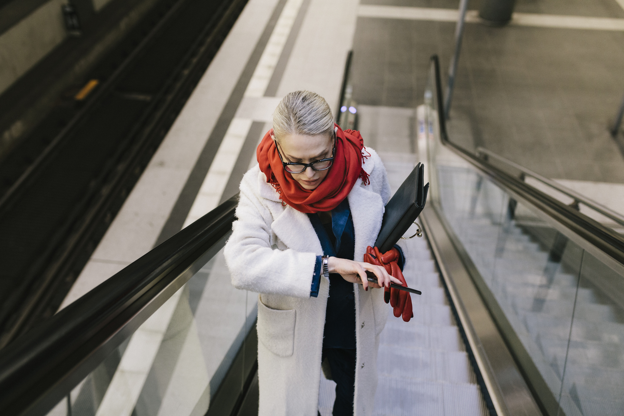 Woman in a white coat using an escalator, looking at her phone, with a red scarf and glasses
