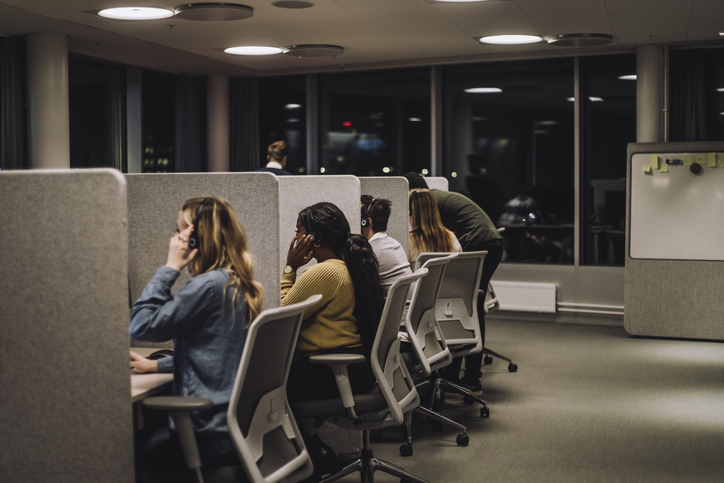 Office workers at desks with headsets, facing computer screens, engaged in customer service or telemarketing