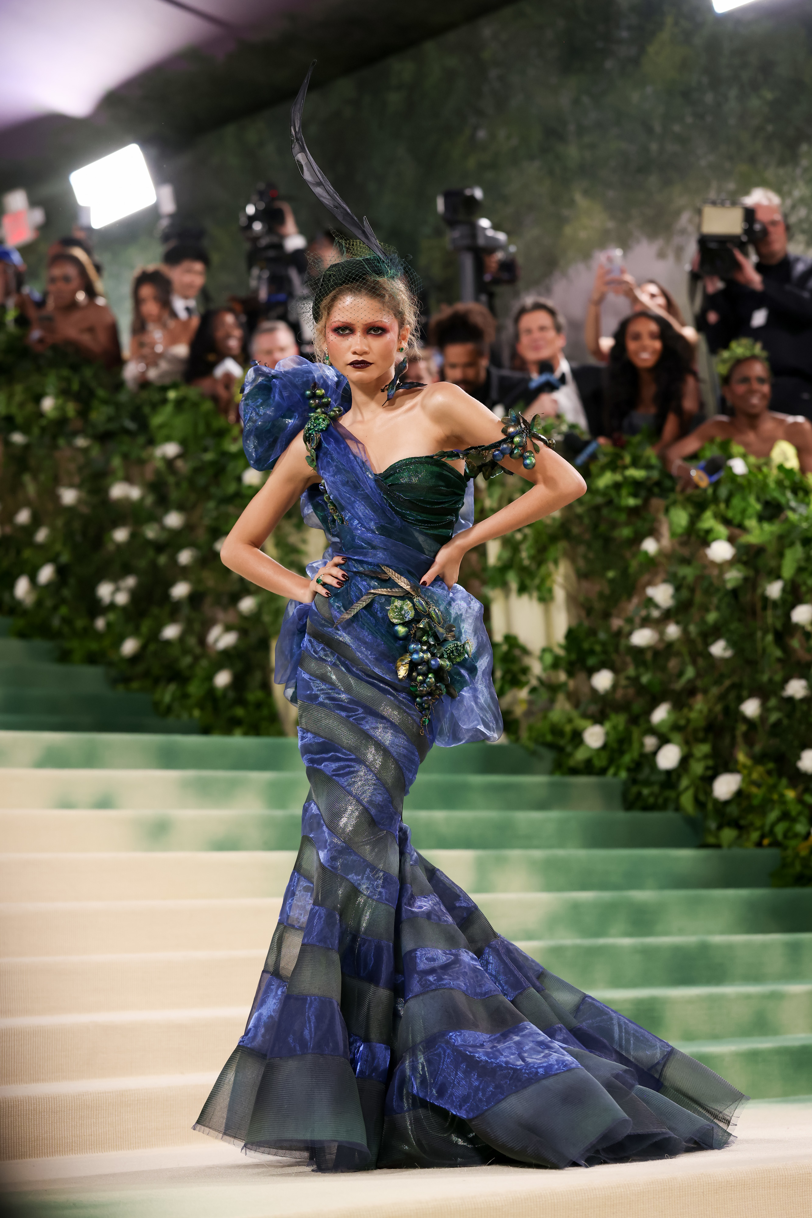 Zendaya in a sculptural blue gown with green accents on a staircase at a gala event, photographers in the background