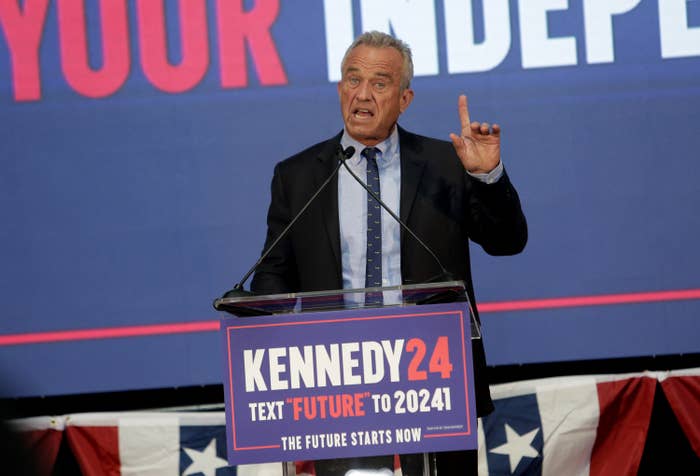 RFK Jr. at podium with &quot;Kennedy 24&quot; sign, gesturing with one hand while speaking