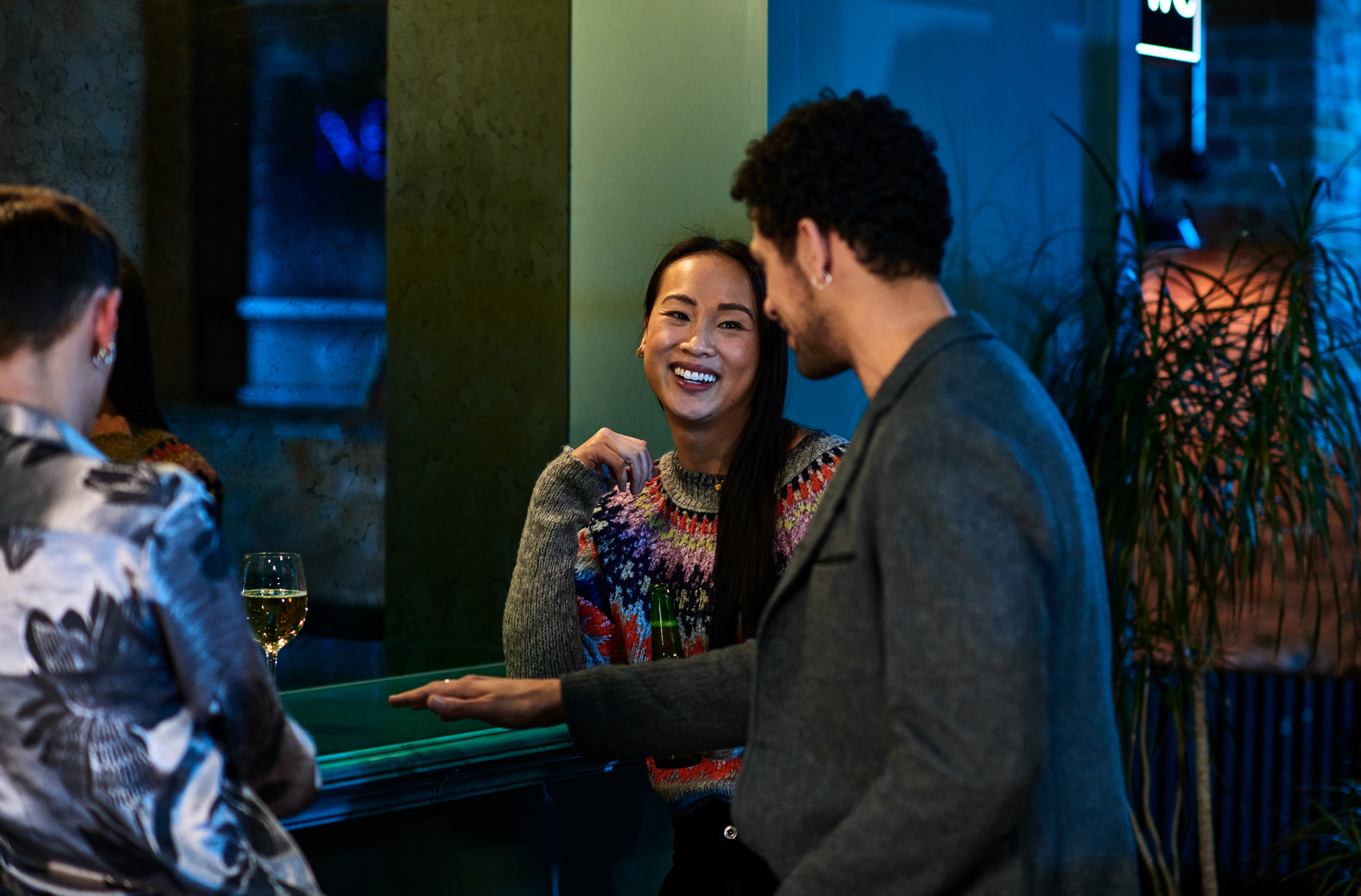 Two people are engaged in a conversation with smiles, in a social setting with a bar in the background
