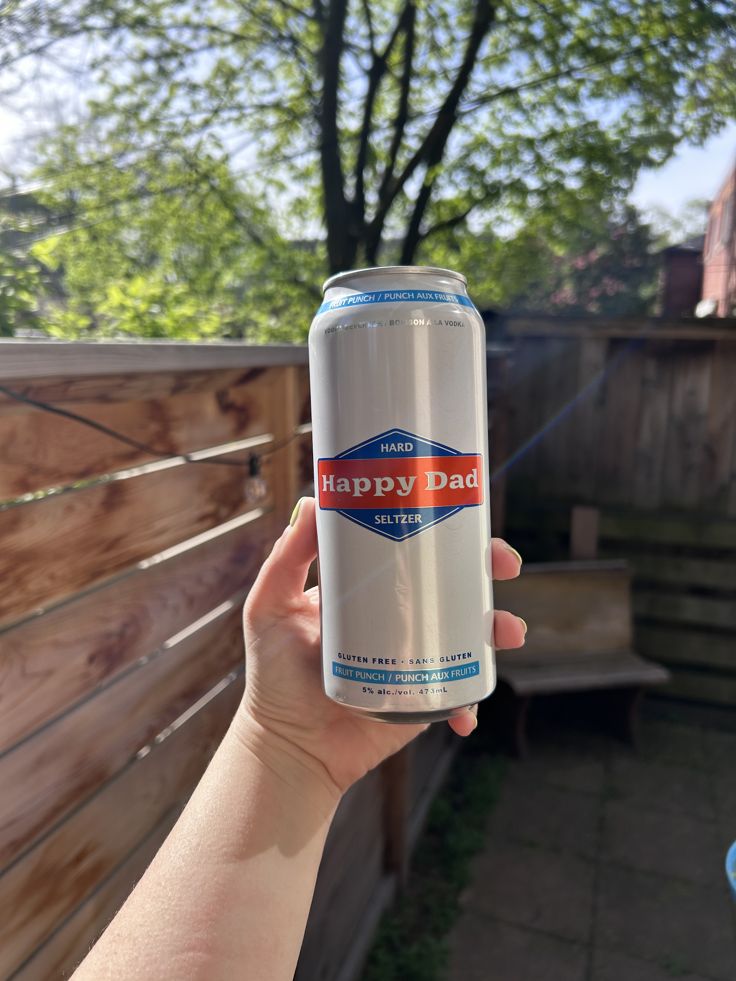 Hand holding a can of Happy Dad hard seltzer outdoors, with foliage and a fence in the background