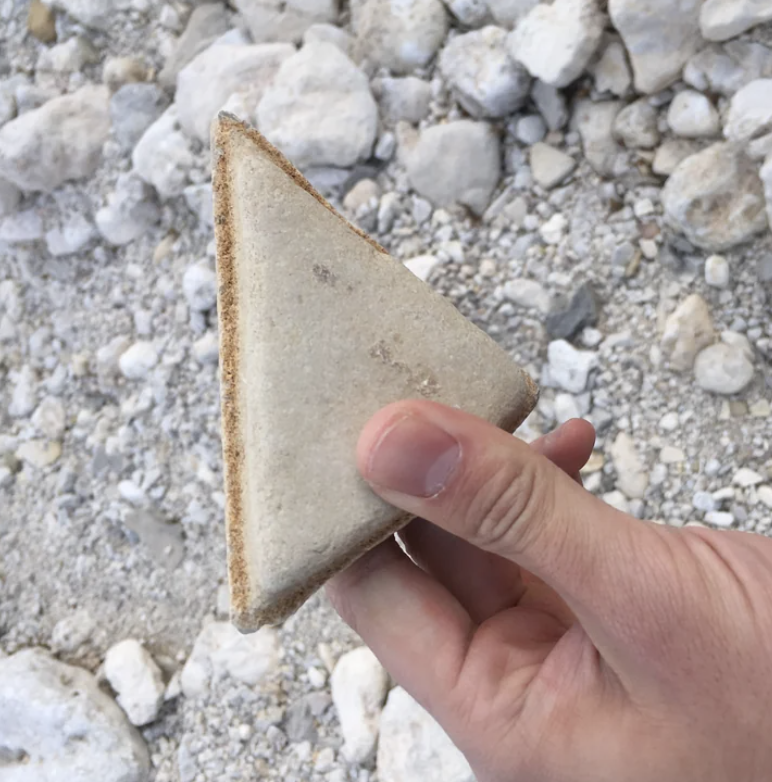 A person holding a triangular rock resembling a sandwich above gravel