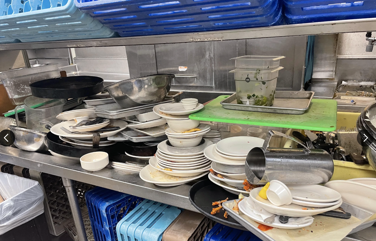 A cluttered commercial kitchen counter stacked with dirty dishes and utensils