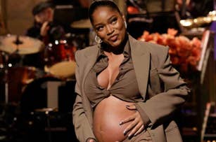 Pregnant celebrity in a tailored suit cradles her belly with a smile on stage, with musicians in the background